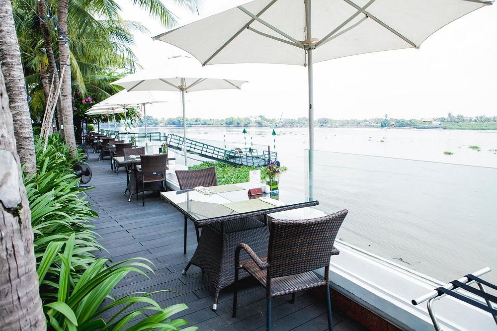 Khám Phá Những Quán Cafe View Sông Sài Gòn Đẹp Nhất Không Thể Bỏ Lỡ 15 Hàng ghế và bàn ngoài trời có mái che, nhìn ra sông, với cây xanh xung quanh và bầu trời sáng.