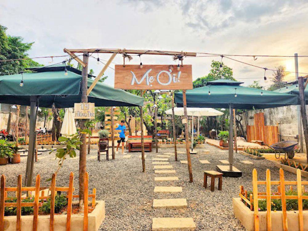 Khám Phá Top 10 Quán Cafe Sân Vườn Đẹp và Thư Giãn Tại Thủ Đức 5 Quán cà phê ngoài trời với biển hiệu, bàn ghế gỗ, ô che màu xanh, cây xanh và lối đi lát đá nhỏ.