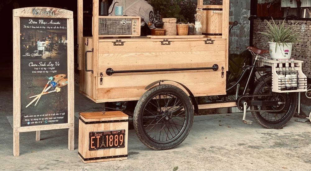 Khám Phá 10 Quán Cafe Gò Vấp Với Góc "Hidden Gem" Độc Đáo 27 Xe đạp bán cà phê với thùng gỗ, bảng menu đứng bên cạnh và chậu cây trang trí.