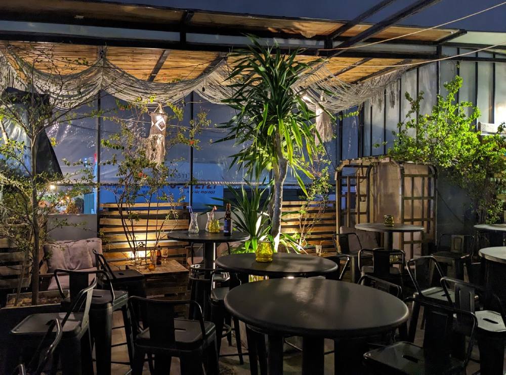 Top 11+ Quán Cafe Đẹp Tại Đà Nẵng Không Thể Bỏ Qua Năm 2025 26 Quán cà phê ngoài trời buổi tối, bàn ghế kim loại, cây xanh trang trí, ánh sáng ấm áp và đồ trang trí treo.