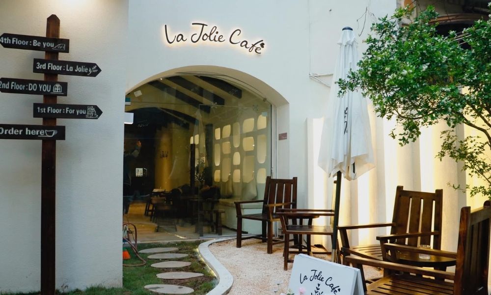 Khám Phá Top Quán Cafe Hấp Dẫn Trên Đường Nguyễn Chí Thanh - Điểm Đến Thú Vị 9 Lối vào quán cà phê với biển chỉ đường và bàn ghế gỗ, cây xanh bên cạnh, ánh sáng ấm áp từ bên trong.