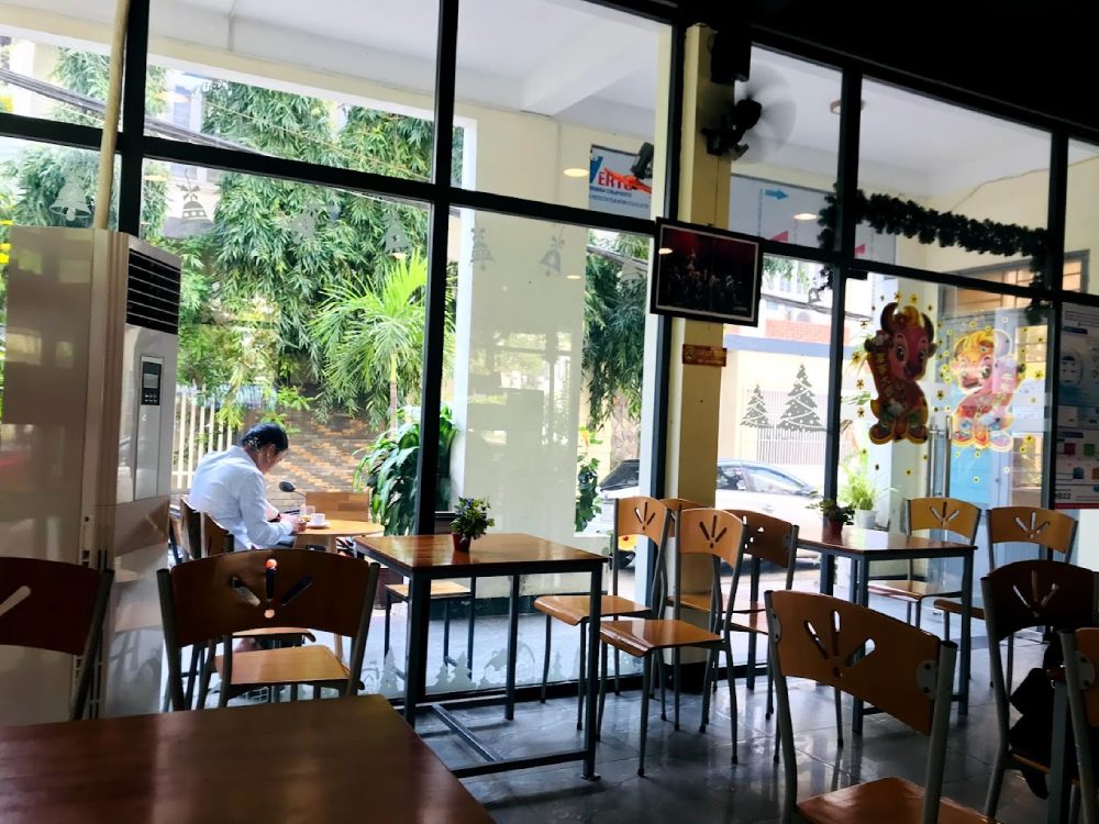 Top 11+ Quán Cafe Đẹp Tại Đà Nẵng Không Thể Bỏ Qua Năm 2025 14 Quán cà phê với bàn ghế gỗ, cửa kính lớn, cây xanh bên ngoài; người ngồi viết tại bàn gần cửa sổ.