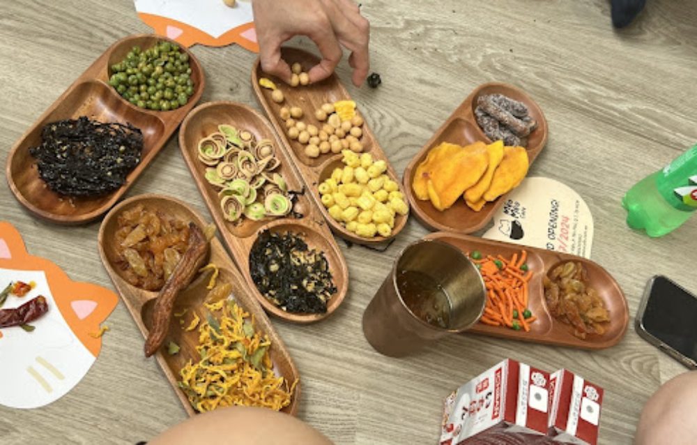 Khám Phá 8 Quán Cafe Mèo TPHCM: Thiên Đường Cho Tín Đồ Yêu Mèo 20 Đĩa gỗ chứa nhiều loại đồ ăn vặt trên sàn, có bàn tay đang lấy thức ăn.