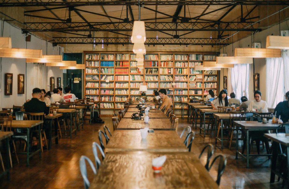 Quán cà phê ấm cúng với kệ sách lớn, ánh sáng tự nhiên, bàn gỗ và khách hàng thư giãn đọc sách.