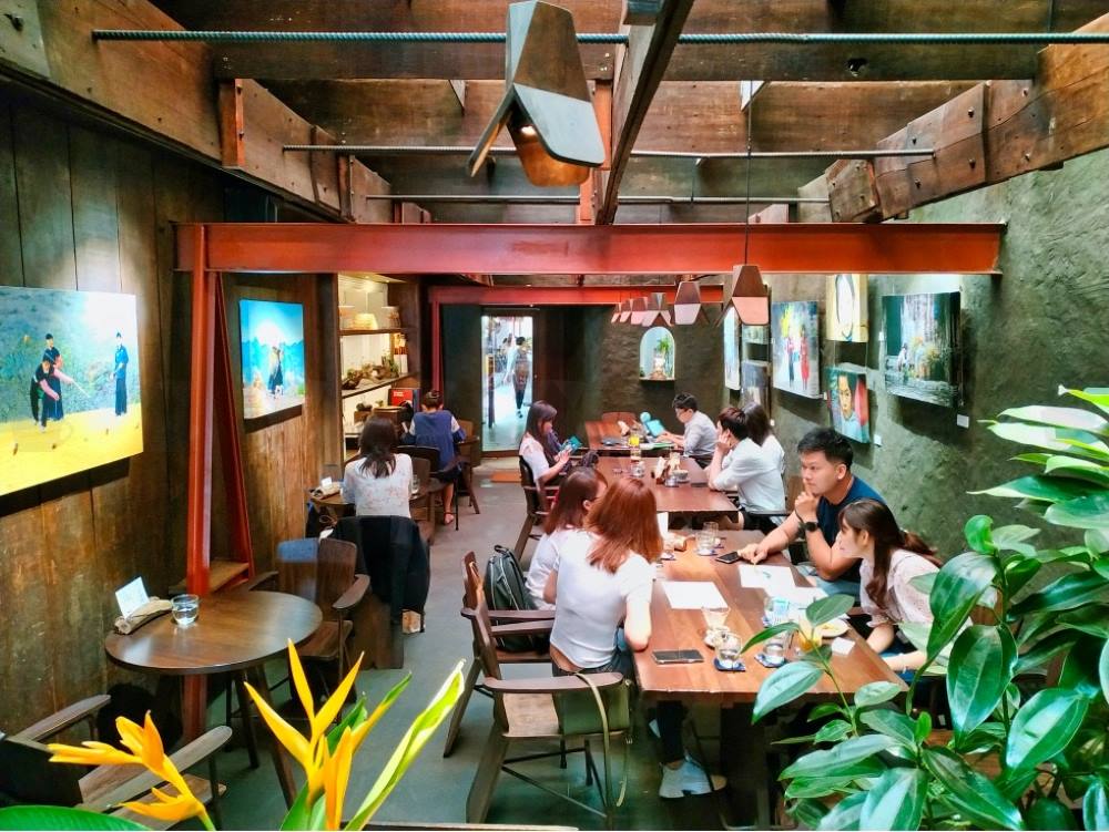 Qu&aacute;n c&agrave; ph&ecirc; với nhiều người ngồi l&agrave;m việc, xung quanh trang tr&iacute; tranh ảnh v&agrave; c&acirc;y xanh.