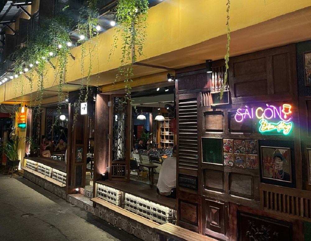 12 Quán Cafe Sài Gòn Xưa Hấp Dẫn Nhất định Phải Thử 5 Quán cà phê với đèn neon và cây xanh trang trí bên ngoài, có khách ngồi bên trong, không gian ấm cúng và thư giãn.