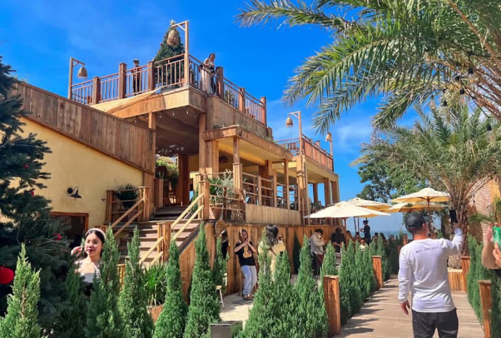Khám Phá 12+ Quán Cafe Ngắm Hoàng Hôn Tuyệt Đẹp Tại Vũng Tàu 9 Quán cà phê ngoài trời, có cầu thang gỗ, cây xanh, ô che nắng và nhiều người tham quan dưới trời nắng.