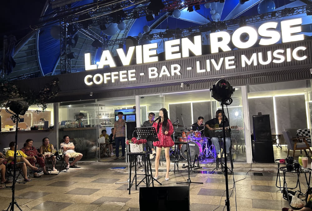 Khám Phá 12+ Quán Cafe Ngắm Hoàng Hôn Tuyệt Đẹp Tại Vũng Tàu 7 Ca sĩ biểu diễn trước quán cà phê có biển hiệu "LA VIE EN ROSE", xung quanh có khán giả ngồi thưởng thức.