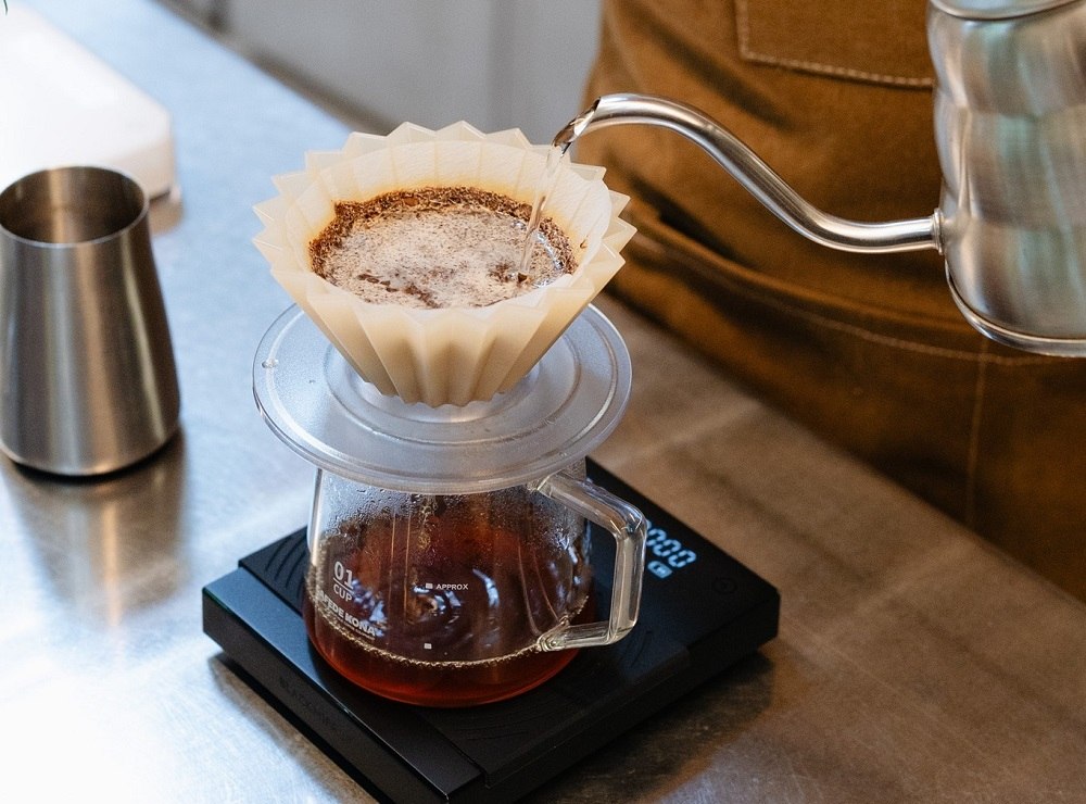 Khám Phá Những Quán Cafe View Sông Sài Gòn Đẹp Nhất Không Thể Bỏ Lỡ 17 Người đang pha cà phê bằng phương pháp pour-over với ấm đun nước và bộ lọc giấy trên cân điện tử.