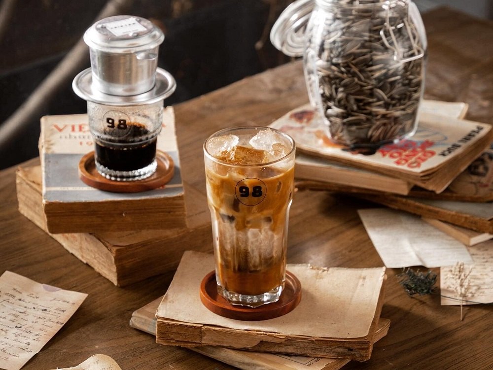 12 Quán Cafe Sài Gòn Xưa Hấp Dẫn Nhất định Phải Thử 11 Ly cà phê sữa đá và cà phê phin trên bàn, bên cạnh sách và lọ hạt hướng dương.