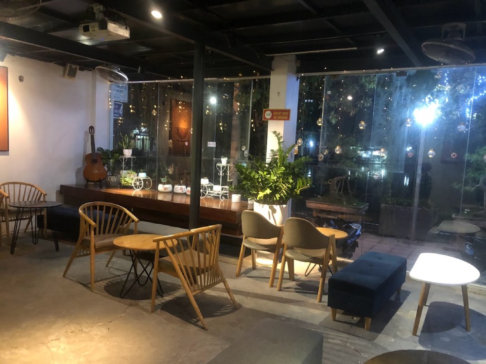 Khám Phá Garden Coffee Đội Cấn: Không Gian Yên Tĩnh Giữa Lòng Thủ Đô 6 Quán cà phê với bàn ghế gỗ, đàn guitar và cây xanh, ánh sáng dịu từ đèn và cửa kính lớn nhìn ra ngoài.
