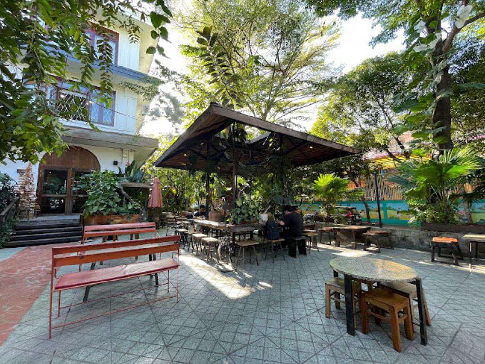 Khám Phá Top 10 Quán Cafe Sân Vườn Đẹp và Thư Giãn Tại Thủ Đức 9 Quán cà phê ngoài trời với bàn ghế gỗ, cây xanh bao quanh, và tòa nhà phía sau vào một ngày nắng.
