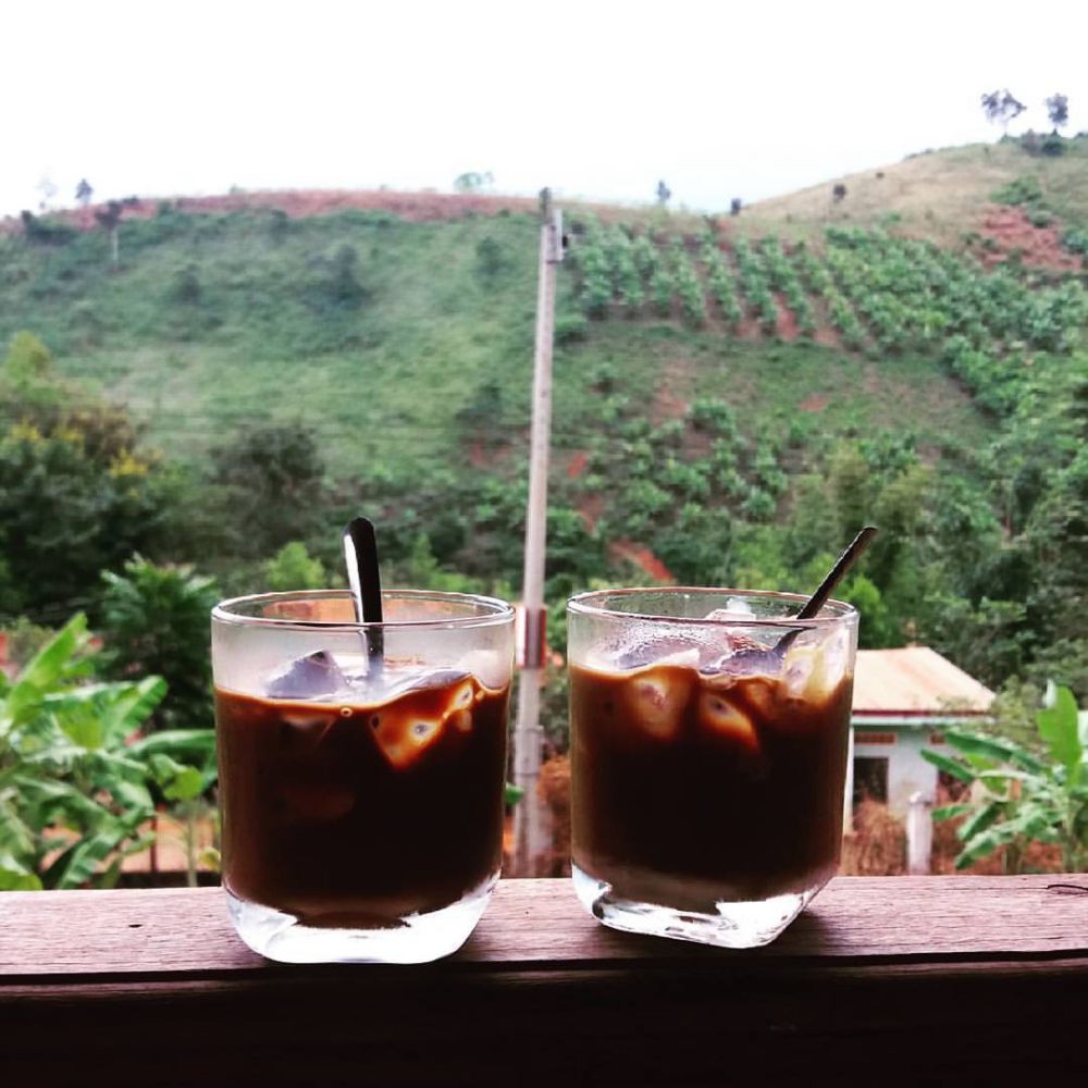 Hai ly cà phê sữa đá trên lan can gỗ, phía sau là đồi núi xanh tươi với cây cối và nhà nhỏ.