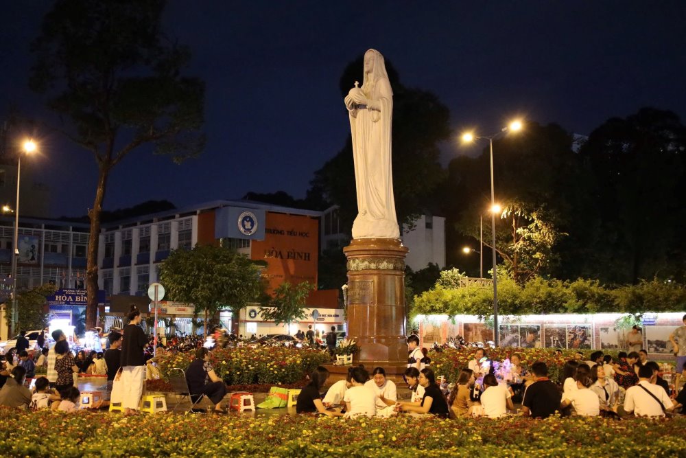 Nhiều người tụ tập buổi tối quanh bức tượng lớn trong c&ocirc;ng vi&ecirc;n, &aacute;nh đ&egrave;n đường chiếu s&aacute;ng khung cảnh xung quanh.