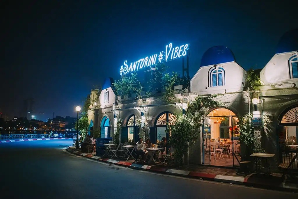 Khám Phá Top 10 Quán Cafe Hồ Tây View Đẹp Ngắm Hoàng Hôn Lãng Mạn 9 Quán cà phê phong cách Santorini vào ban đêm với ánh đèn neon và cây cối trang trí, có khách ngồi bên ngoài.