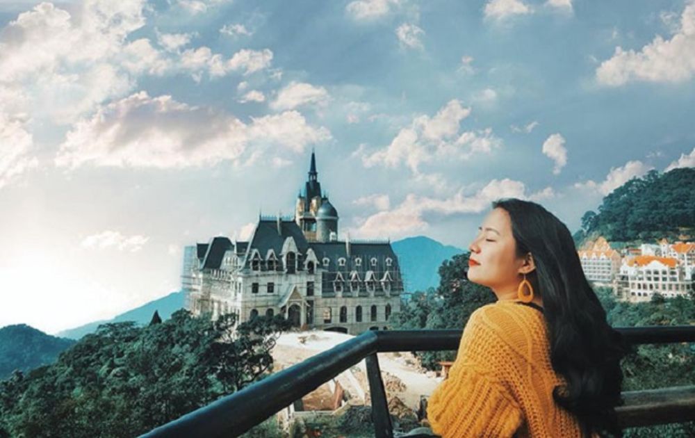 Khám Phá Quán Gió Tam Đảo: Trải Nghiệm Cafe Trên Mây Với View Tuyệt Đẹp 6 Người đứng trên ban công, phía sau là tòa lâu đài và bầu trời xanh có mây trắng.