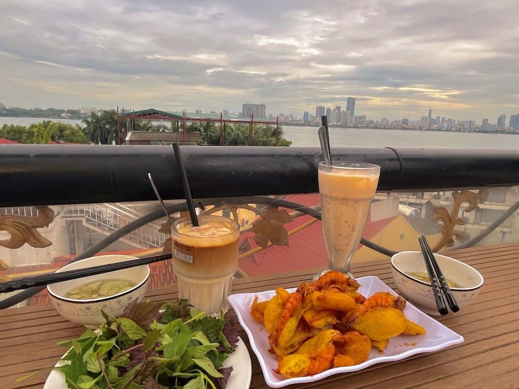 Khám Phá Top 10 Quán Cafe Hồ Tây View Đẹp Ngắm Hoàng Hôn Lãng Mạn 4 Bàn ăn ngoài trời với đồ ăn và thức uống, nhìn ra cảnh thành phố và sông dưới bầu trời nhiều mây.