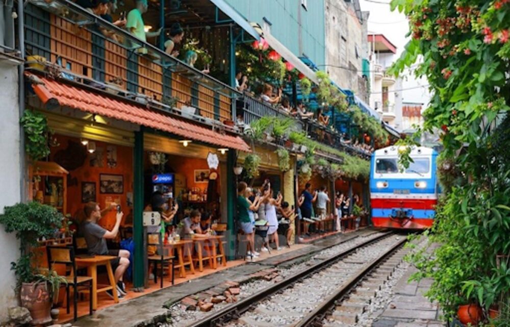 Khám Phá Phố Đường Tàu Phùng Hưng: Trải Nghiệm Cà Phê Độc Đáo Tại Hà Nội 6 Đoàn tàu chạy qua phố nhỏ với quán cà phê hai bên, nhiều người đứng xem và chụp ảnh.