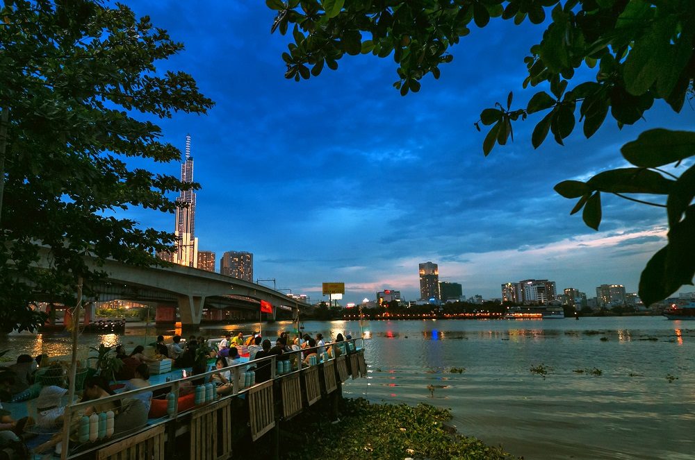 Khám Phá Những Quán Cafe View Sông Sài Gòn Đẹp Nhất Không Thể Bỏ Lỡ 22 Khung cảnh thành phố về đêm với tòa nhà cao tầng, cầu và sông, người dân ngồi thư giãn bên bờ sông.