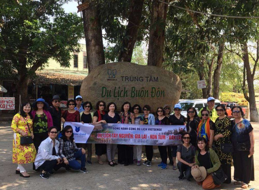 Một nhóm người đứng trước tảng đá có chữ "Trung Tâm Du Lịch Buôn Đôn", cầm băng rôn du lịch.