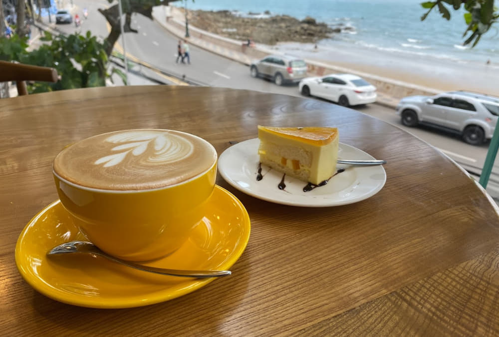 Khám Phá 12+ Quán Cafe Ngắm Hoàng Hôn Tuyệt Đẹp Tại Vũng Tàu 5 Ly cà phê và bánh ngọt trên bàn gỗ nhìn ra đường ven biển với xe cộ và biển xanh.