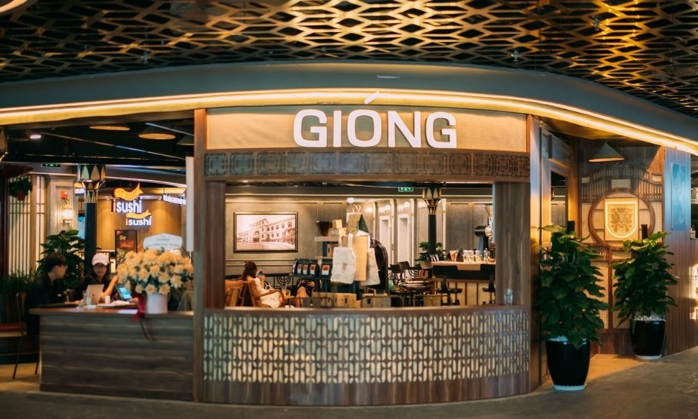 Khám Phá 15 Quán Cafe Yên Tĩnh ở Quận 1: Không Gian Thoải Mái, Thức Uống Tuyệt Hảo 19 Mặt tiền quán cà phê với bảng hiệu lớn, không gian nội thất sang trọng, có cây xanh trang trí và khách ngồi bên trong.