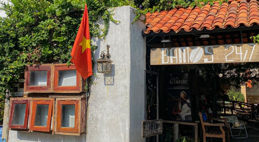 Khám Phá 10 Quán Cafe Gò Vấp Với Góc "Hidden Gem" Độc Đáo 21 Quán cà phê nhỏ với mái ngói đỏ, cửa sổ gỗ, cờ đỏ sao vàng, bảng hiệu "Bamos 24/7", cây xanh bao quanh.