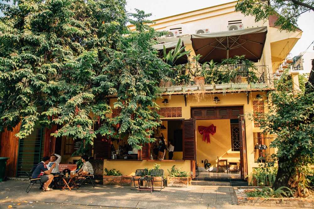 12 Quán Cafe Sài Gòn Xưa Hấp Dẫn Nhất định Phải Thử 18 Quán cà phê với cây xanh bao quanh, có người ngồi bên ngoài, mặt tiền trang trí đơn giản và ấm cúng.