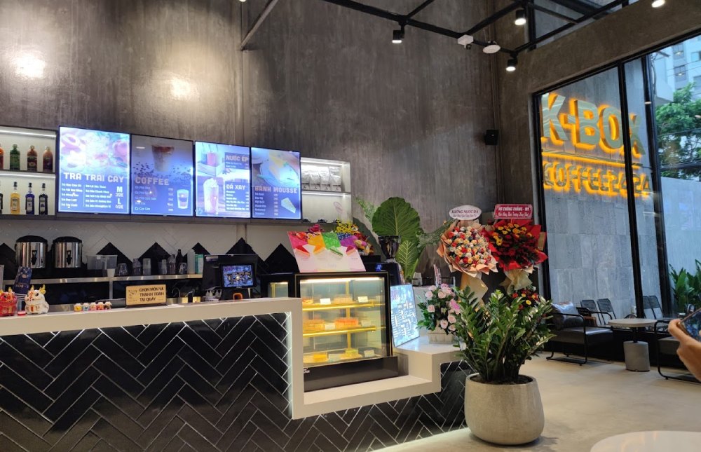 Khám Phá 8+ Quán Cafe Đêm Sài Gòn Đẹp Lung Linh Bạn Không Thể Bỏ Lỡ 17 Quán cà phê hiện đại với quầy bar, thực đơn trên màn hình, bánh ngọt trong tủ kính và nhiều chậu hoa trang trí.