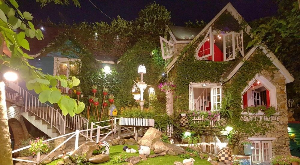 Khám Phá 10 Quán Cafe Gò Vấp Với Góc "Hidden Gem" Độc Đáo 30 Ngôi nhà đá phủ cây xanh, có cửa sổ mở, xung quanh là vườn hoa và đèn sáng vào ban đêm.