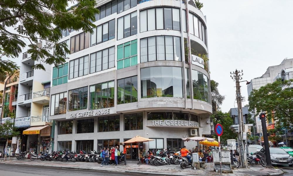 Khám Phá 15 Quán Cafe Yên Tĩnh ở Quận 1: Không Gian Thoải Mái, Thức Uống Tuyệt Hảo 29 Tòa nhà hiện đại bên góc đường, nhiều xe máy đậu phía trước, có quán cà phê và người đi bộ.