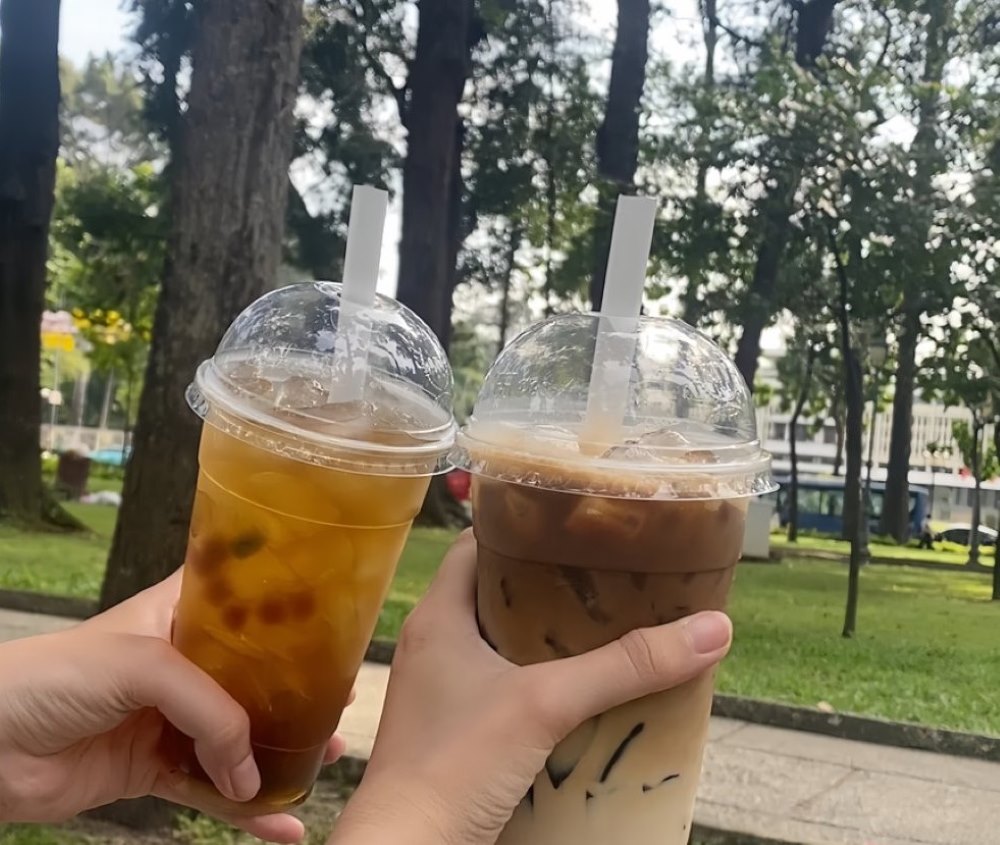 Hai ly tr&agrave; sữa đang được n&acirc;ng l&ecirc;n trong c&ocirc;ng vi&ecirc;n xanh, nền l&agrave; h&agrave;ng c&acirc;y v&agrave; b&atilde;i cỏ.