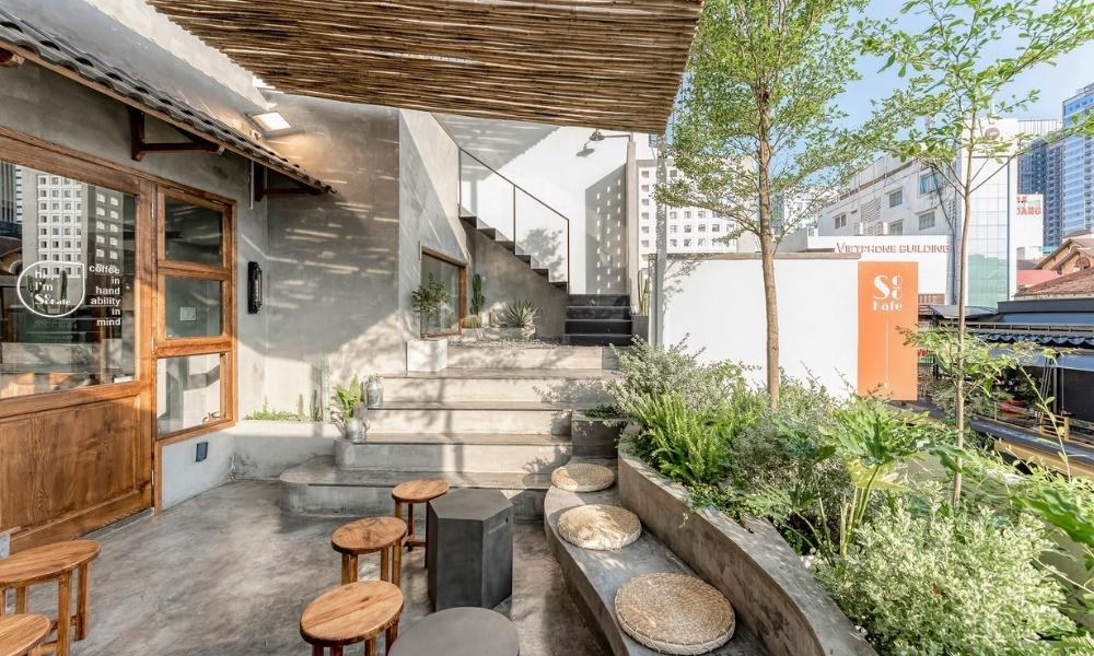 Khám Phá 15 Quán Cafe Yên Tĩnh ở Quận 1: Không Gian Thoải Mái, Thức Uống Tuyệt Hảo 7 Khu vực ngoài trời của quán cà phê với bàn ghế gỗ và cây xanh, cầu thang dẫn lên tầng trên, ánh sáng tự nhiên.