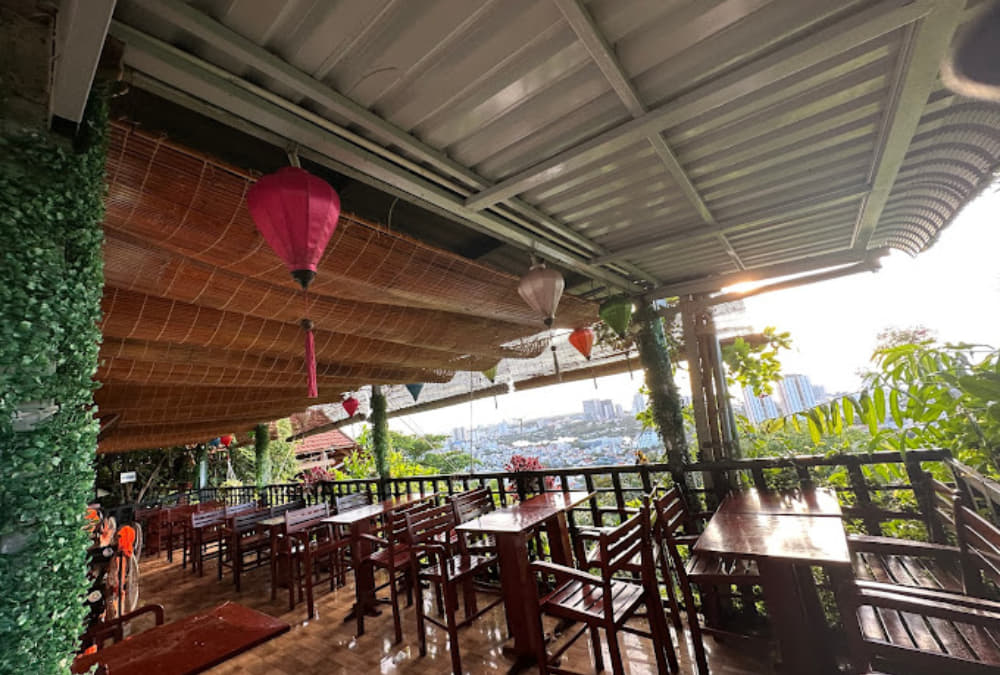 Khám Phá 12+ Quán Cafe Ngắm Hoàng Hôn Tuyệt Đẹp Tại Vũng Tàu 8 Quán cà phê ngoài trời với bàn ghế gỗ, đèn lồng treo, cây xanh xung quanh và tầm nhìn ra thành phố.