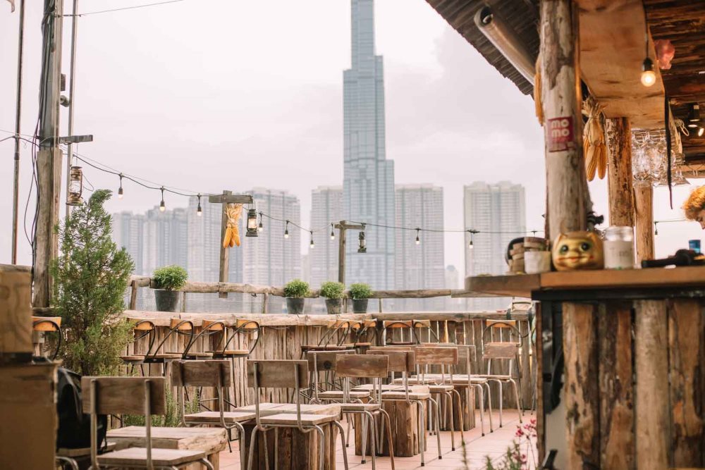 Qu&aacute;n c&agrave; ph&ecirc; ngo&agrave;i trời với b&agrave;n ghế gỗ, đ&egrave;n treo v&agrave; t&ograve;a nh&agrave; cao tầng ph&iacute;a xa trong khung cảnh đ&ocirc; thị.