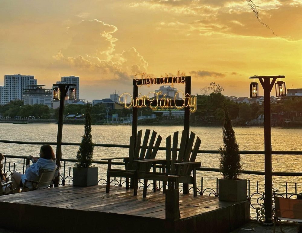 Qu&aacute;n c&agrave; ph&ecirc; ven s&ocirc;ng l&uacute;c ho&agrave;ng h&ocirc;n, với ghế gỗ v&agrave; bảng hiệu đ&egrave;n neon, khung cảnh th&agrave;nh phố ph&iacute;a xa.