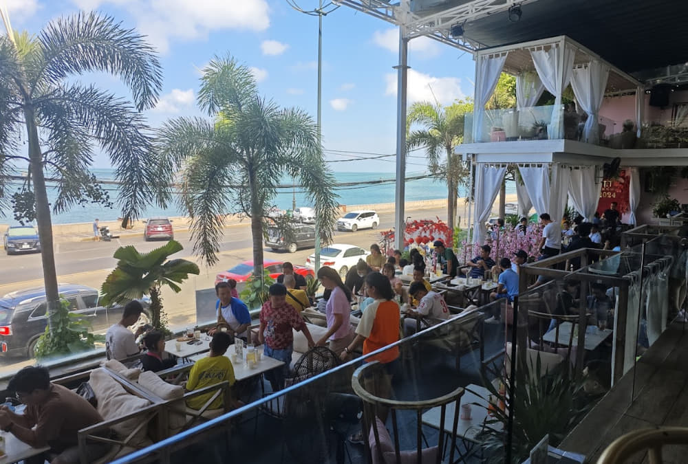 Khám Phá 12+ Quán Cafe Ngắm Hoàng Hôn Tuyệt Đẹp Tại Vũng Tàu 6 Quán cà phê ven biển, nhiều người ngồi ăn uống, khung cảnh biển xanh và hàng dừa phía sau.