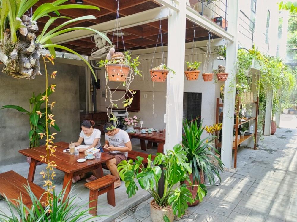 Hai người ngồi ăn uống tại b&agrave;n gỗ ngo&agrave;i trời, xung quanh c&oacute; nhiều c&acirc;y xanh v&agrave; chậu hoa treo.