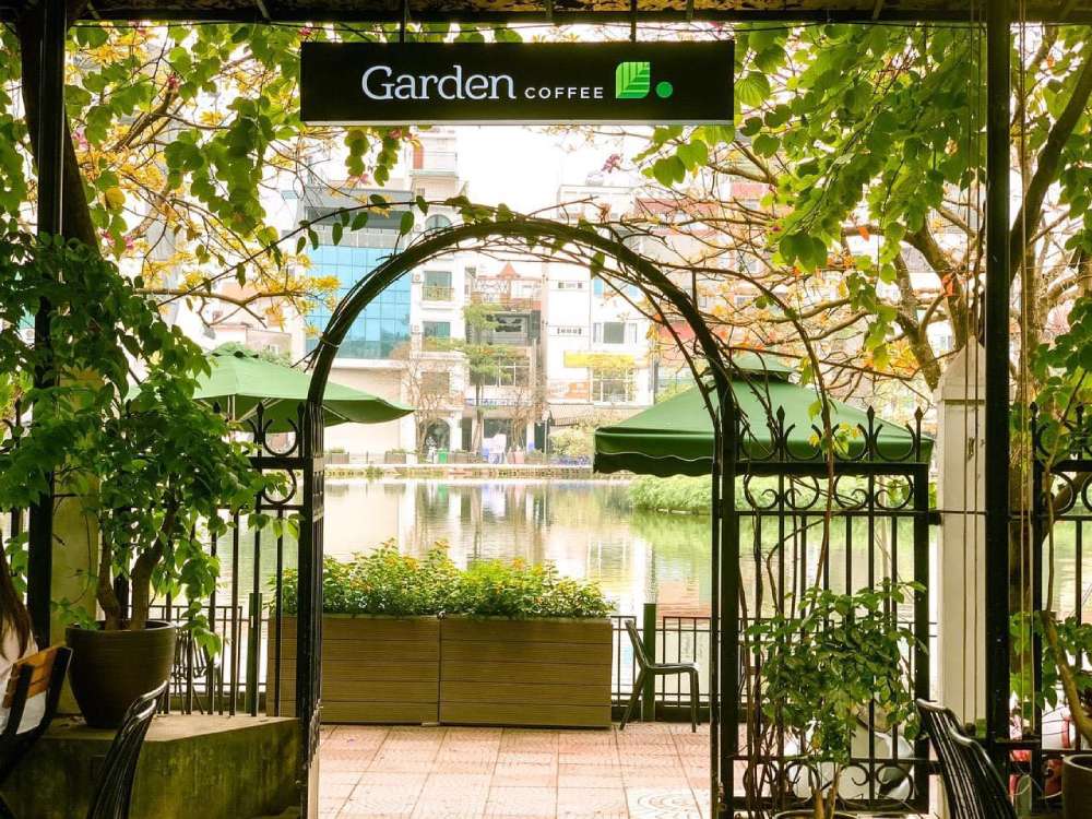 Khám Phá Garden Coffee Đội Cấn: Không Gian Yên Tĩnh Giữa Lòng Thủ Đô 3 Cổng vòm sắt dẫn vào quán cà phê bên hồ, có cây xanh và dù che, tạo không gian thoáng đãng, yên bình.