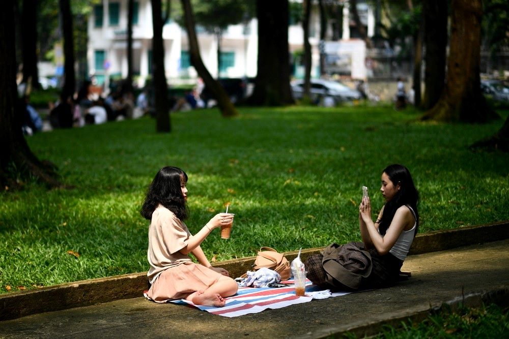 Khám Phá Top 6+ Quán Cà Phê Vỉa Hè Sài Gòn Được Giới Trẻ Yêu Thích 5 Hai người ngồi trên thảm picnic ở công viên, một người chụp ảnh, người kia cầm ly nước.