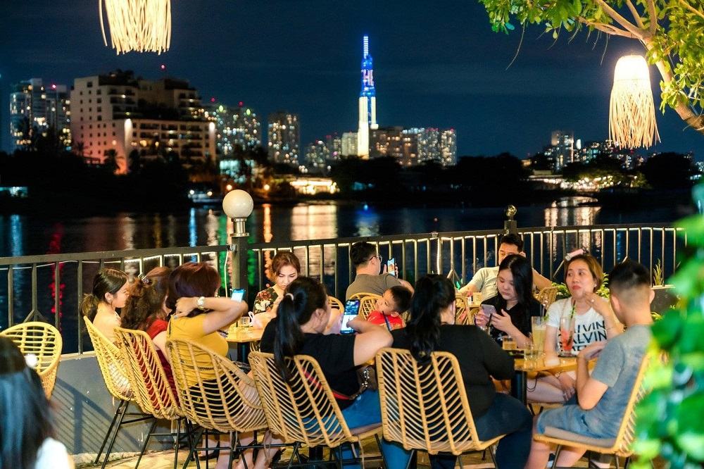 Khám Phá Những Quán Cafe View Sông Sài Gòn Đẹp Nhất Không Thể Bỏ Lỡ 11 Nhóm người ngồi ăn uống tại quán cà phê ven sông vào buổi tối, với cảnh thành phố sáng đèn phía xa.