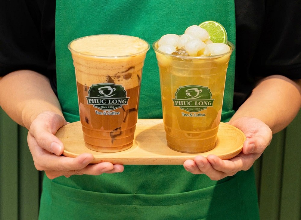 Khám Phá Những Quán Cafe View Sông Sài Gòn Đẹp Nhất Không Thể Bỏ Lỡ 19 Hai ly đồ uống trên khay gỗ, một ly có lớp bọt, ly còn lại có đá và lát chanh, được cầm bởi người mặc tạp dề xanh.