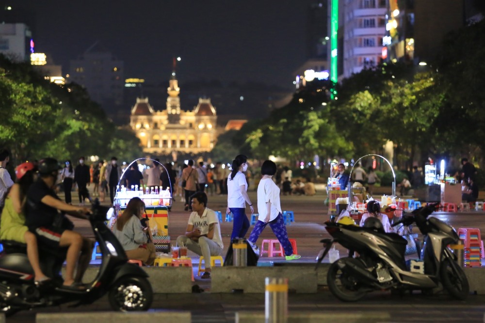 Đường phố về đ&ecirc;m với người đi bộ, xe m&aacute;y, v&agrave; quầy h&agrave;ng nhỏ; t&ograve;a nh&agrave; chiếu s&aacute;ng ph&iacute;a xa.