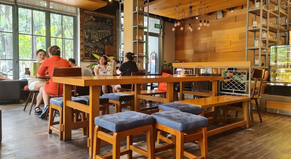 Khám Phá 8+ Quán Cafe Đêm Sài Gòn Đẹp Lung Linh Bạn Không Thể Bỏ Lỡ 7 Quán cà phê với bàn ghế gỗ, ánh sáng tự nhiên từ cửa sổ, và một số người ngồi trò chuyện.