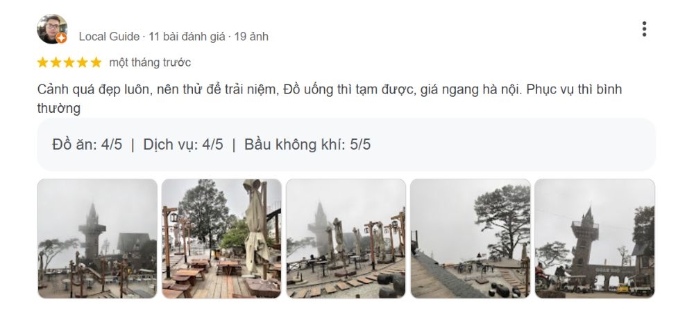 Khám Phá Quán Gió Tam Đảo: Trải Nghiệm Cafe Trên Mây Với View Tuyệt Đẹp 9 Quán cà phê ngoài trời với bàn ghế gỗ, cây cối và tượng trong không gian sương mù, có cổng chào và tòa tháp phía xa.