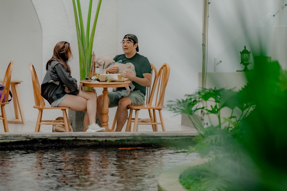 Hai người ngồi đối diện nhau tại b&agrave;n c&agrave; ph&ecirc; ngo&agrave;i trời, c&oacute; c&acirc;y xanh v&agrave; hồ nước b&ecirc;n cạnh.