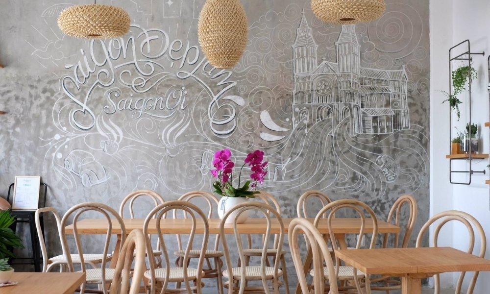 Khám Phá 15 Quán Cafe Yên Tĩnh ở Quận 1: Không Gian Thoải Mái, Thức Uống Tuyệt Hảo 17 Quán cà phê với tường vẽ phấn nhà thờ và dòng chữ nghệ thuật, bàn ghế gỗ và chậu hoa trang trí.