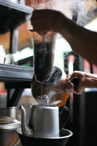 Người pha c&agrave; ph&ecirc; bằng c&aacute;ch r&oacute;t nước qua phin lọc, hơi nước bốc l&ecirc;n từ ấm kim loại.