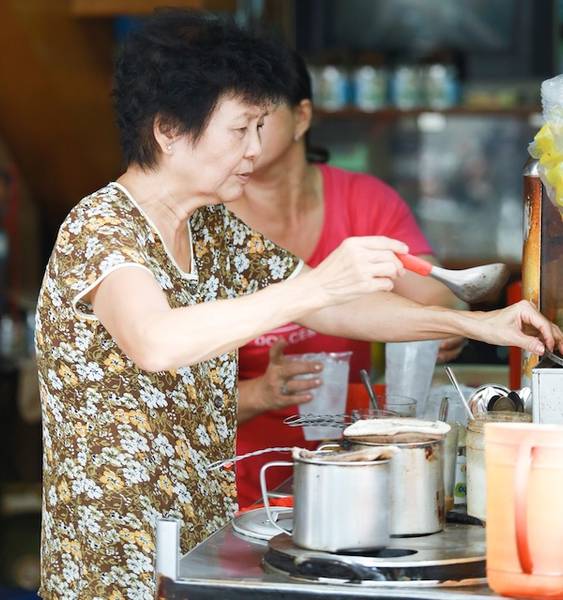 Người phụ nữ mặc &aacute;o hoa đang pha chế đồ uống tại quầy, xung quanh c&oacute; nhiều dụng cụ v&agrave; chai lọ.