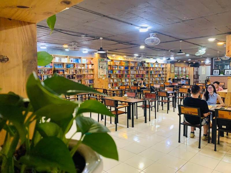 cafe-sach-ha-noi-1-1024x768-1 Quán cà phê với bàn ghế gỗ, kệ sách đầy sách, cây xanh ở góc trái, hai người ngồi trò chuyện.
